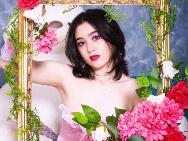 Annakomatsu, met zwart haar, roze oogmake-up en rode lippenstift, wordt omlijst door een gouden spiegel met roze, witte en rode bloemen en groene wijnranken, en toont haar borsten..