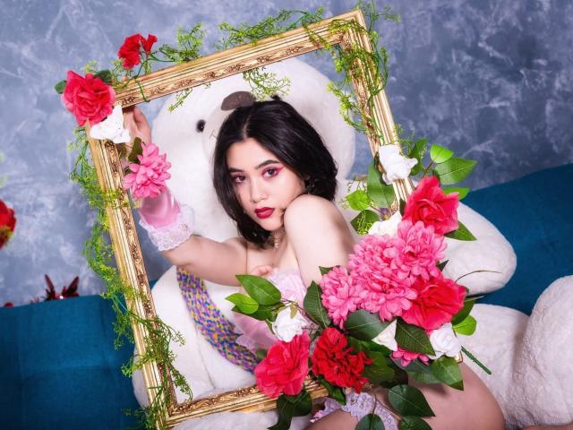 Annakomatsu met een roze bloemenlijst, witte kant en rode rozen, zittend op een blauwe bank, een lijst vasthoudend, een groot wit pluche dier, donker golvend haar, rode lippenstift en roze oogmake-up..