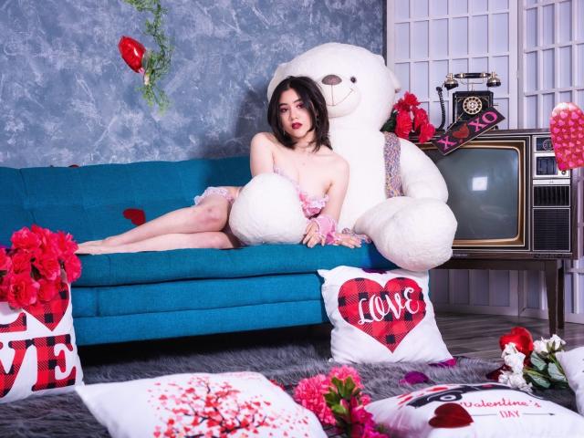 Annakomatsu, avec sa peau pâle, ses cheveux noirs et sa lingerie rose, est assise sur un canapé bleu, avec un grand ours en peluche blanc, des ballons rouges et une décoration de Saint-Valentin, montrant ses fesses, entourée de fleurs..