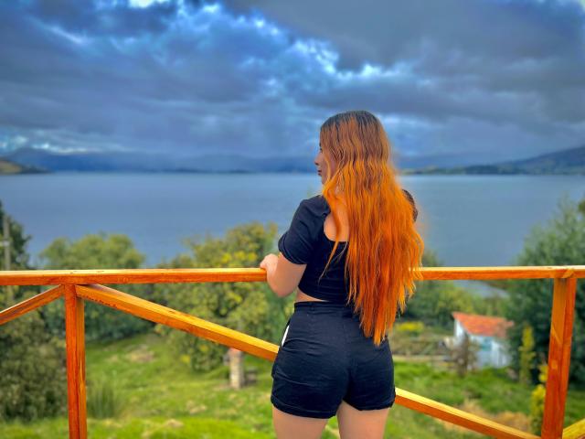 Daniela, una donna con lunghi capelli arancioni, una maglietta nera e pantaloncini neri, in piedi su un balcone di legno, di fronte a un lago blu, sotto un cielo nuvoloso. Le sue natiche sono leggermente visibili..