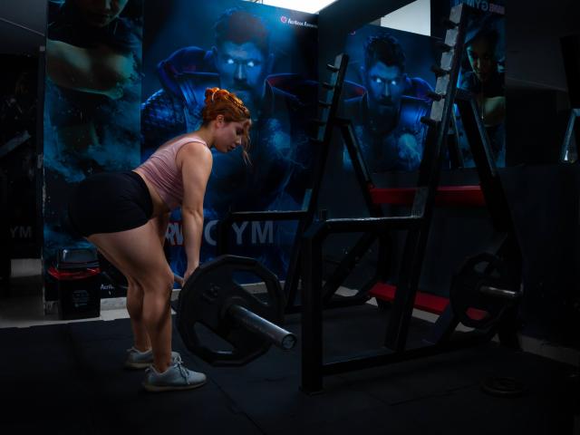 DanielaSiren with red hair, pink tank top, black shorts, white sneakers, bending over a weight bench, focusing on a barbell, in a dimly lit gym.