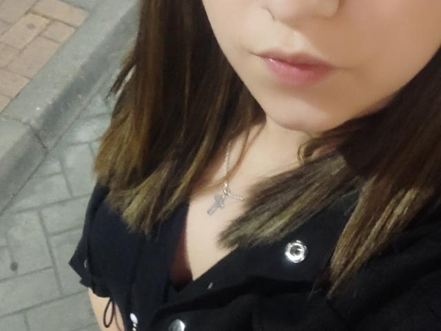 Kkatalina's face with slightly parted pink lips, brown shoulder-length hair, black button-up shirt, and silver necklace with cross pendant. Boobs visible under shirt. Upper chest focus.