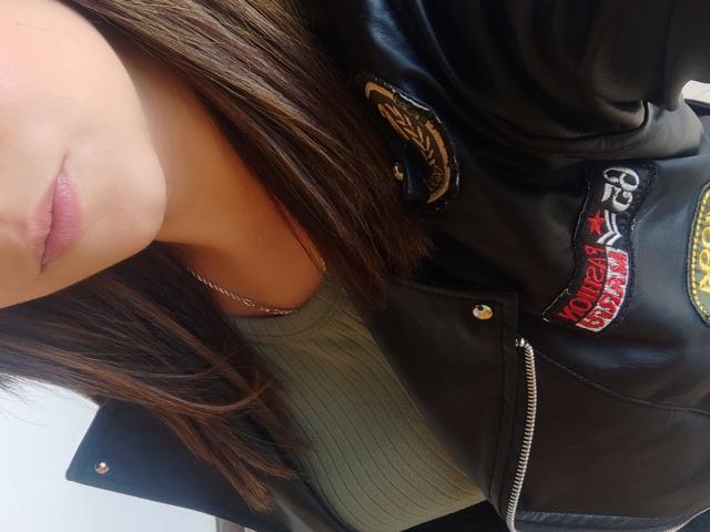 Kkatalina's face with light pink lips, wearing a black leather jacket with a "5-10" patch, over a green ribbed top, and a silver necklace, brown hair in frame.