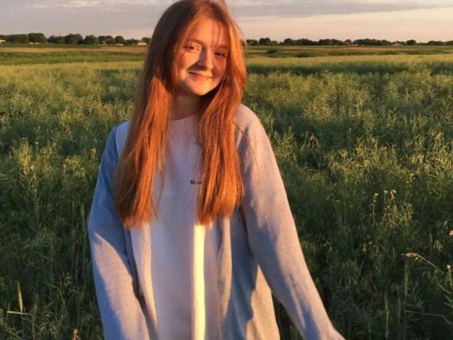 LaPochka, com longos cabelos ruivos, sorrindo, vestindo uma camiseta branca e um cardigã azul, está em pé em um campo verde durante o pôr do sol. Seus seios são pequenos e ela é fofa..