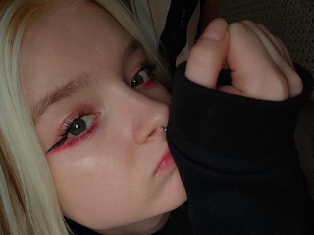 LaPochka has pale skin, blonde hair, and pink eyeliner with black winged edges. She's biting her black sleeve with a nose piercing and slightly parted lips.