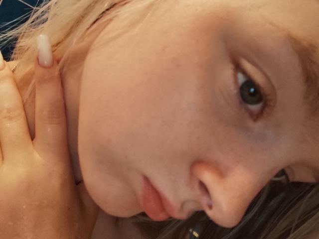 LaPochka's face, showing her pale skin, brown eyes, and pink lips, with a hand gently touching her neck. Blonde hair, slightly messy, and a small nose piercing visible.