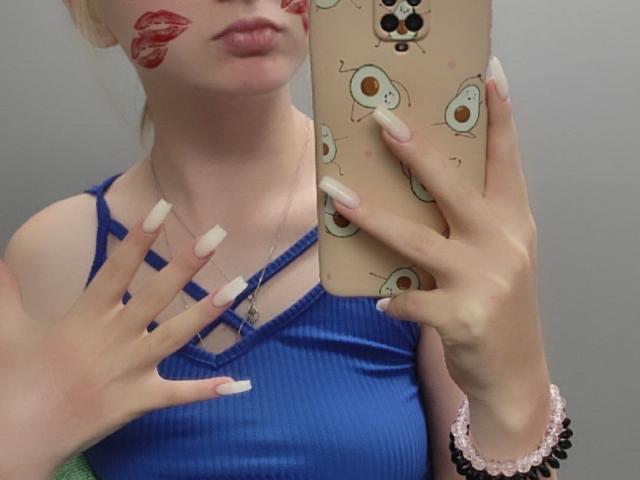 LaPochka with red lipstick marks on her cheeks, wearing a blue ribbed tank top, holding a phone with egg-themed cover, long white nails, pink bracelet, mirror selfie.