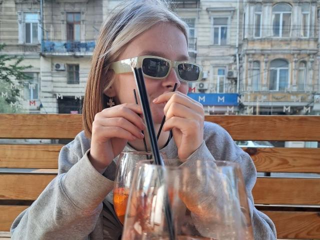LaPochka with blonde hair, wearing sunglasses, grey hoodie, drinking from a glass with a black straw, holding a second glass, wooden bench in the background.