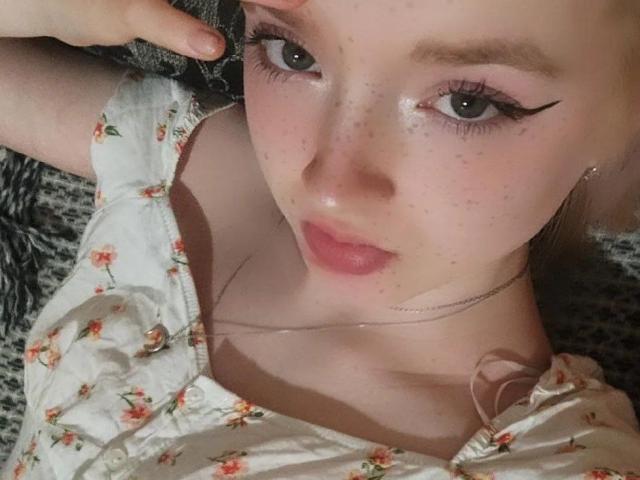 El rostro de LaPochka, con pecas, lápiz labial rosa y delineador de ojos negro. Lleva un vestido floral, está acostada y tocándose la cabeza. Sus senos son ligeramente visibles..