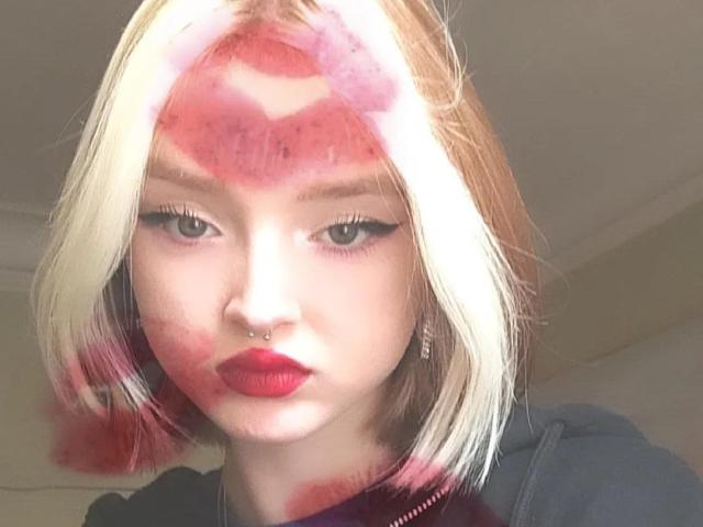 LaPochka mit rotem Lippenstift-Kuss auf der Stirn, Nasenpiercing, rotem Lippenstift, blonden und pinken Haaren, Eyeliner, die in die Kamera schaut, trägt ein dunkles Oberteil, hat helle Haut, ist süß..