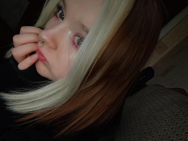 LaPochka with blonde and brown ombre hair, nose piercing, and pink eyeshadow, biting her finger, grey eyes, black top, hand near mouth. 