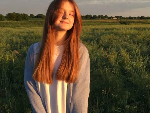 LaPochka, com longos cabelos ruivos, olhos fechados e um sorriso, vestindo um moletom claro azul e branco, está em um campo gramado ao pôr do sol..