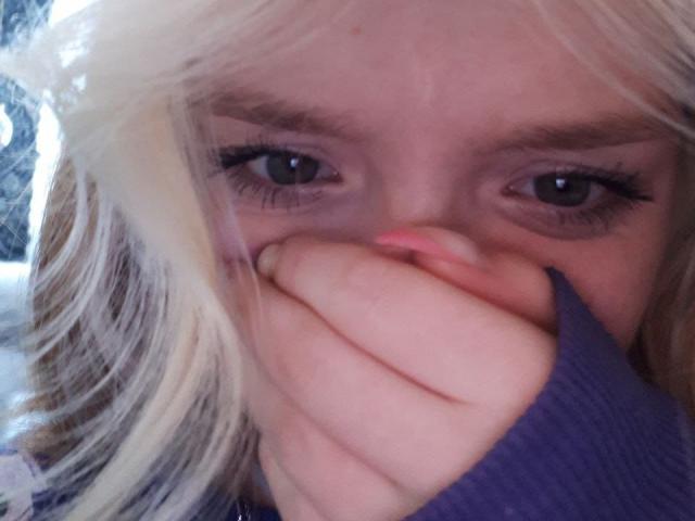 LaPochka mit blonden Haaren, blauen Augen und langen Wimpern, die Nase und Mund mit einem violetten Finger bedecken, und die leicht zur Seite schaut. Sie trägt einen gerippten, violetten Pullover..