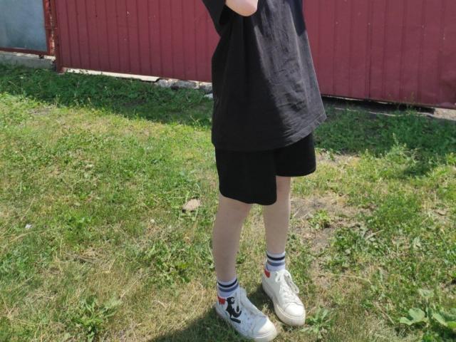 LaPochka, mit schwarzem T-Shirt, schwarzen Shorts, weißen Socken mit Streifen und weißen Turnschuhen, steht auf grünem Gras, im Hintergrund ist eine rote Holzwand zu sehen..