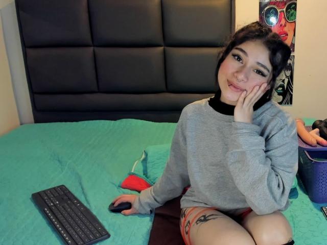 Hesttia, with dark hair and eyeliner, sits on a bed in a grey sweater, resting her cheek on her hand, with a keyboard and mouse on the green quilt.