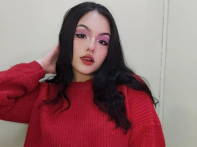 HaleiBeckham with long black hair, red ribbed sweater, pink eye makeup, and parted lips, hand in hair, looking slightly to the side, pale skin, sexy, cute.