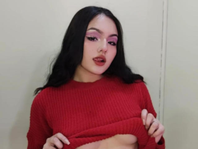 HaleiBeckham with long black hair, pink eyeshadow, and red ribbed sweater, lifting it to expose her small tits and flat stomach. She has a slight, sexy, confident look.