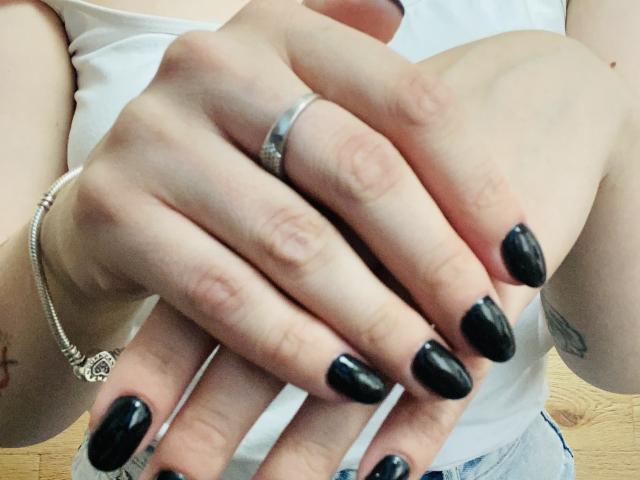 KarKutie's hands with black glossy nails, wearing a silver ring and bracelet, fingers slightly curled, white tank top, light skin, wooden floor, subtle tattoo on wrist.