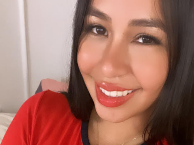 Het gezicht van MeganWisse, met glanzende rode lippenstift, lachend met witte tanden, een rode blouse aan, zwart haar en bruine ogen, ziet er schattig en aantrekkelijk uit..