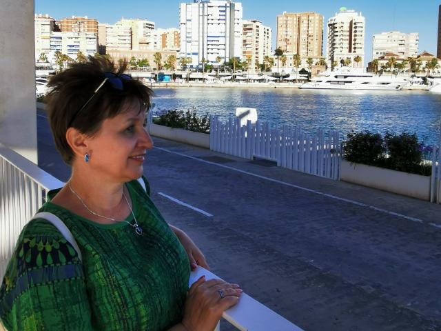 DoraBlueEyess, middle-aged woman with short brown hair, green patterned dress, blue earrings, standing by a white railing, holding a white paper, looking at a waterfront with boats.