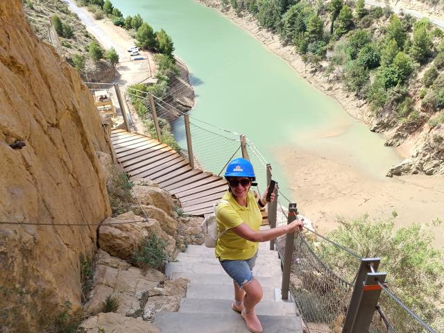 DoraBlueEyess, wearing a blue helmet and yellow shirt, climbs a rocky, metal-cabled staircase with a hand on the railing, looking down at a green river.