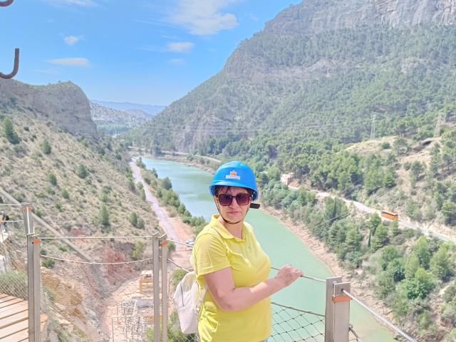 DoraBlueEyess with blue cap, yellow shirt, and sunglasses stands on a rocky bridge, holding a rope, with a green river and mountain valley in the background.