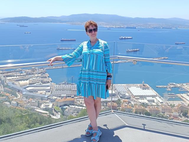 DoraBlueEyess has short brown hair, wears sunglasses, a blue striped dress, and blue sandals, standing on a rooftop with a glass railing, looking over a blue ocean.