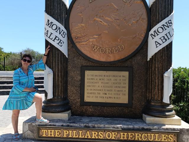 DoraBlueEyess stands with a cute ass in a blue dress, sunglasses, and striped pattern, posing next to a bronze monument with columns and flags, in bright daylight.