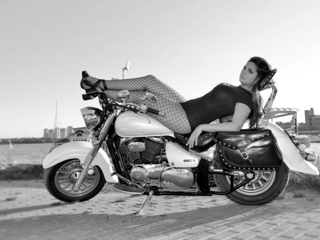 Black-and-white photo of AlinaMilf with fishnet pants, black top, and high heels, lounging on a Harley-Davidson motorcycle, legs up, looking seductive, urban background.