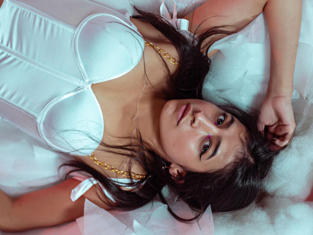 NaiaSanders with her tits in a white, shiny, busty, corset-like top, lying on a fluffy, white surface, arms above her head, brown hair, red eyes, and a slight smile.