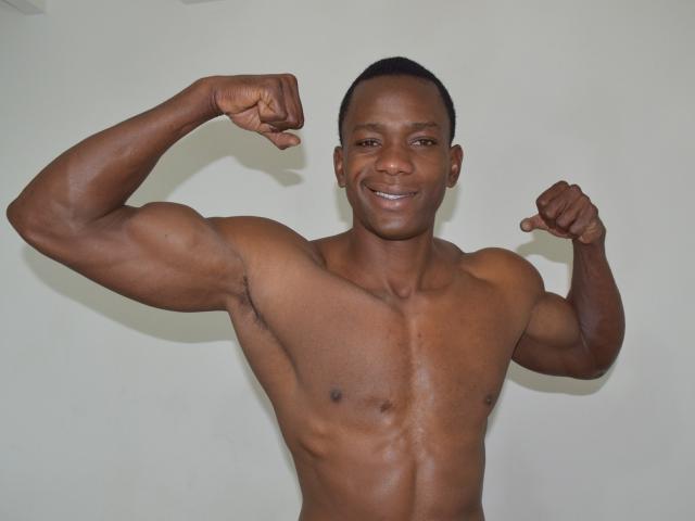 AntonyCruze, un homme musclé à la peau foncée, contracte ses deux bras, montrant ses biceps forts et sa poitrine bien dessinée, il sourit avec confiance, il a les cheveux noirs courts, il est nu et se tient devant un mur blanc uni..