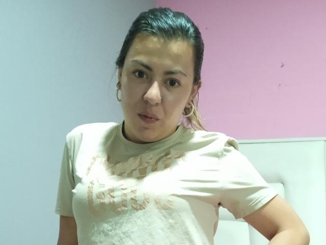 BellaTinya has brown eyes, small earrings, and a light pink t-shirt with a faded design. She's looking slightly to the side with a neutral expression.