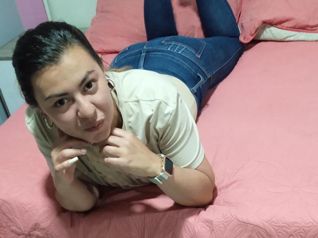 BellaTinya, con cabello oscuro, acostada en una cama rosa, vestida con una camisa beige, jeans azules y un reloj de pulsera, mirando a la cámara con una ligera sonrisa..