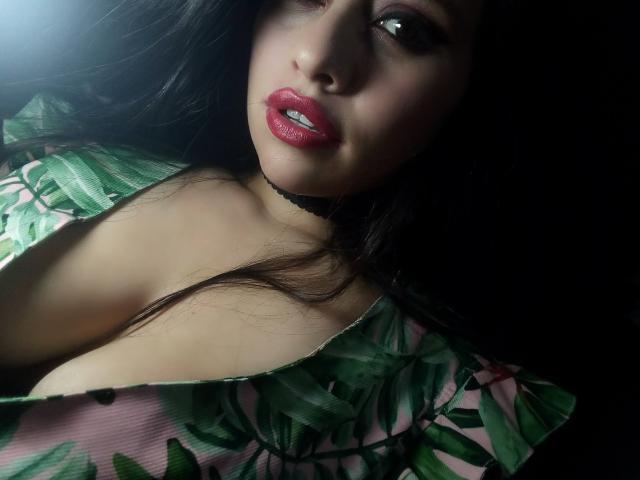BellaSmithX69 heeft glanzende rode lippen, donker golvend haar en een zwarte choker, en draagt een groene top met een bladpatroon, waarbij haar grote borsten zichtbaar zijn, alles in zacht licht..
