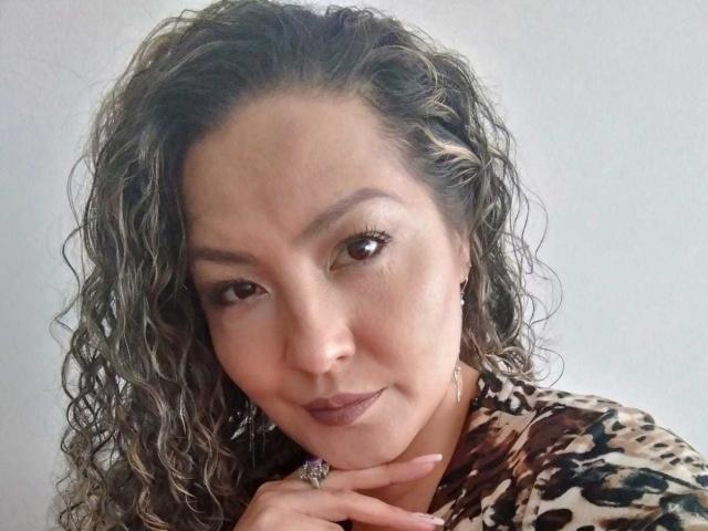Amandabella69 ha i capelli castano ricci, gli occhi castani e un leggero sorriso. Indossa una camicia a stampa leopardo, ha un anello al dito e una posa delle mani rilassata..