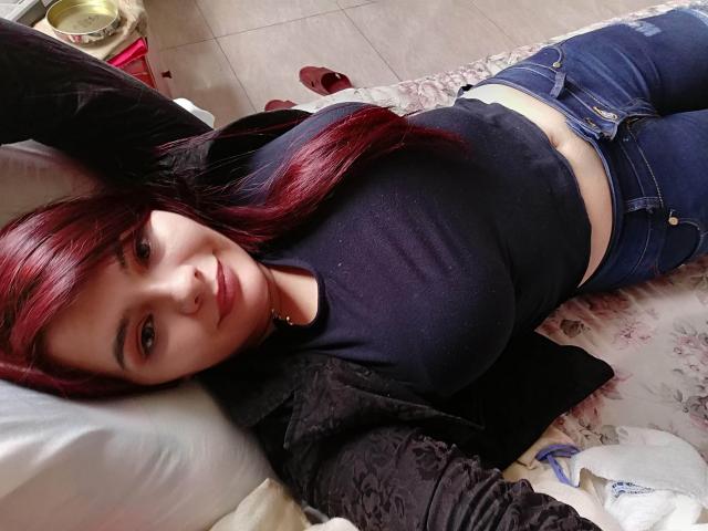KatherinLatin is lying on a bed, wearing a black top and blue jeans, with red hair and a nose ring, showing her tits and a small belly button.