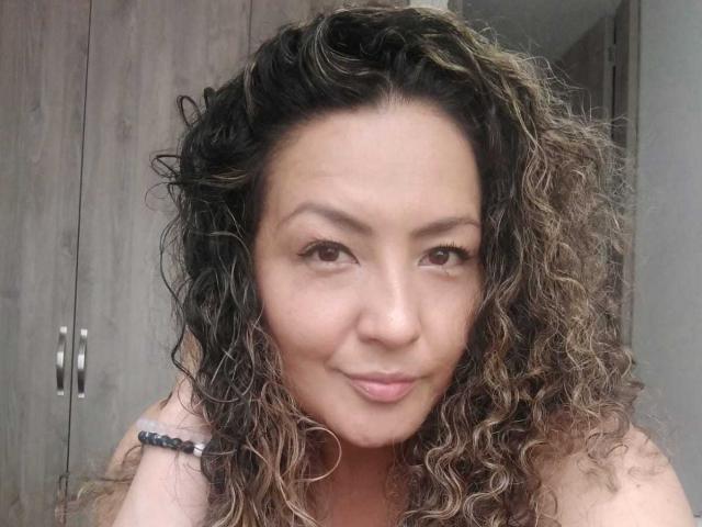 Amandabella69 met krullend bruin en blond haar, lichtbruine huid en een lichte glimlach, waarbij haar gezicht en bovenste schouders zichtbaar zijn. Ze draagt een zwarte kralenarmband om haar linkerpols..