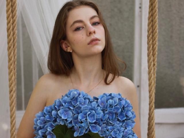 AriaMiss with brown hair, holding a bouquet of blue hydrangeas, has small tits and wears a gold necklace, looking slightly annoyed, in a rope swing.