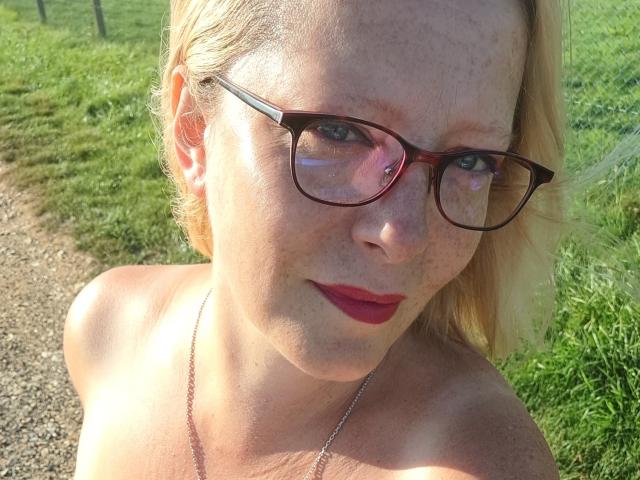 EleanaRose has pale skin, blonde hair, red lipstick, and wears brown glasses. She's topless, showing her bare shoulders and small, freckled breasts. She's outside in sunlight.
