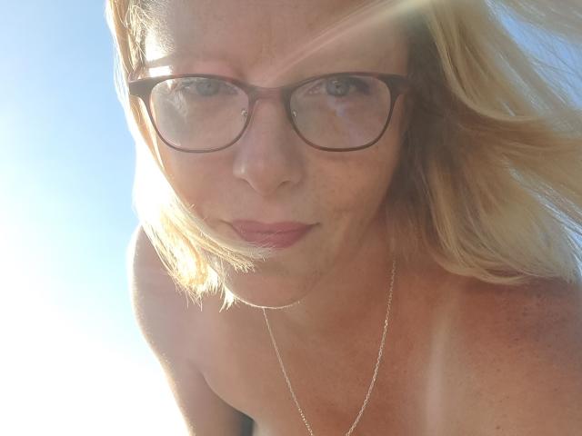 EleanaRose a les cheveux blonds, porte des lunettes et a une expression sérieuse, avec un teint légèrement bronzé par le soleil. Elle porte un collier en argent et est nue, dévoilant ses petites seins fermes..