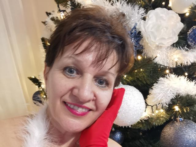 DoraBlueEyess with short brown hair, blue eyes, pink lipstick, smiling, holding red glove, wearing white furry collar, standing in front of decorated Christmas tree with white and blue ornaments.