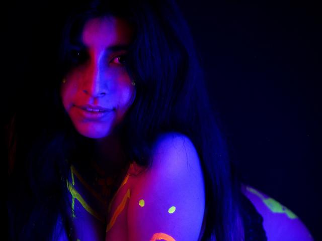 EmmaXWilliams with glowing neon lights on her face and shoulders, smiling, dark wavy hair, blue and purple hues, visible boobs, neon green and pink spots, dark background.