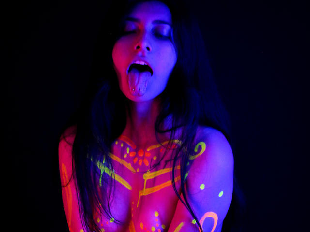 EmmaXWilliams has neon body paint, sticking out her tongue, with glowing yellow and pink patterns on her tits and chest, against a dark background, in a vividly lit photo.