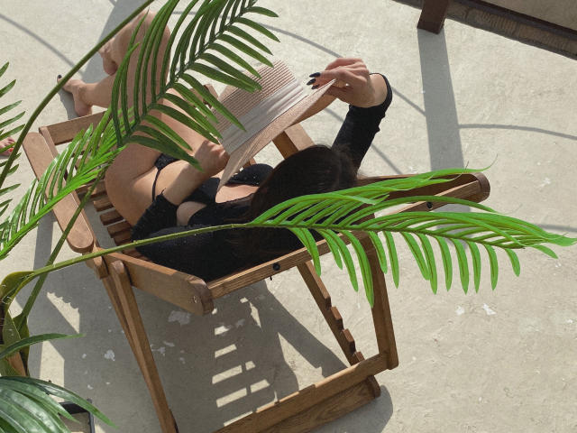 Pingli lounges in a wooden chair, wearing black lingerie, with green palm leaves in front, sun casting shadows, Pingli's hand covers her mouth, black nails, relaxed, sexy.