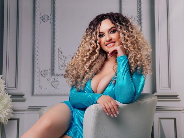 MiaMckenna, avec de grandes seins, vêtue d'une robe bleue brillante, assise sur une chaise, souriante, une main sur la joue, cheveux blonds bouclés, sexy, confiante, attirante, bien éclairée, en intérieur, élégante..