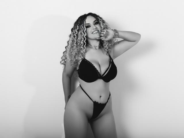 MiaMckenna, con cabello rubio rizado, un sujetador de encaje negro y una tanga a juego, de pie, con la mano en la mejilla, sonriendo, con senos grandes, cintura delgada, sexy, con una pulsera en la muñeca derecha, en blanco y negro..
