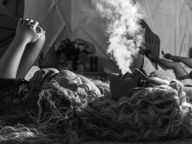MiaMckenna's curly blonde hair, lying on her back, smoking with a large, white plume of smoke, hands up, eyes closed, in a black lace top, in a black-and-white photo.
