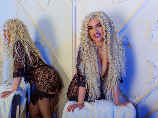 MiaMckenna met lang, krullend blond haar, gekleed in een zwarte kanten robe, die haar achterwerk laat zien, staat met haar handen op een witte stoel, reflectie in een spiegel, blauw licht..