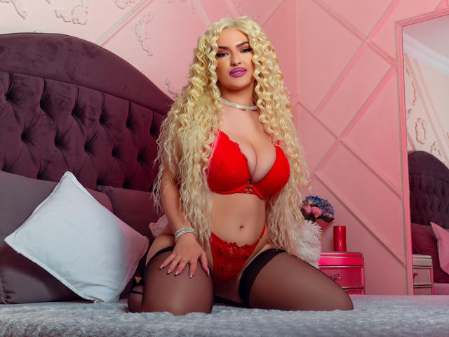 MiaMckenna, con rizos rubios largos, vestida con lencería de encaje roja y medias negras, arrodillada en una cama, mostrando sus grandes senos y su cintura pequeña, en un dormitorio iluminado con luz rosa..