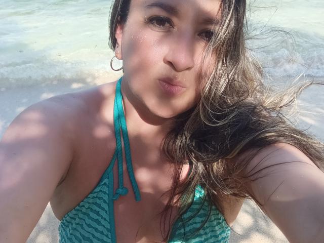 Charlize, con cabello castaño, un bikini azul verdoso a rayas, pequeños aros, labios fruncidos y piel húmeda, de pie en aguas poco profundas, mirando a la cámara, con la luz del sol en el rostro..